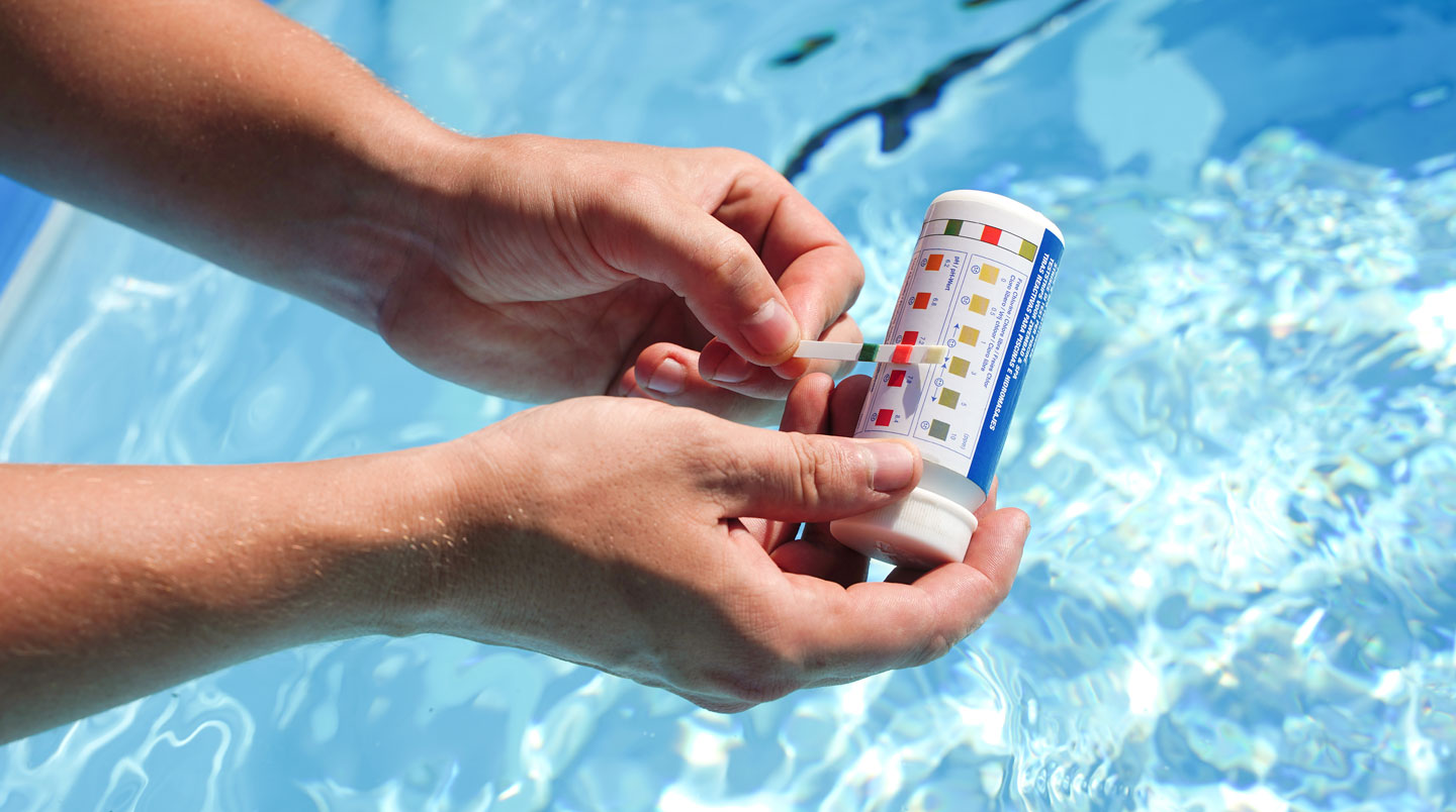 Main plongeant une bandelette de test colorimétrique dans l'eau de piscine avec kit de produits chimiques sur la margelle.